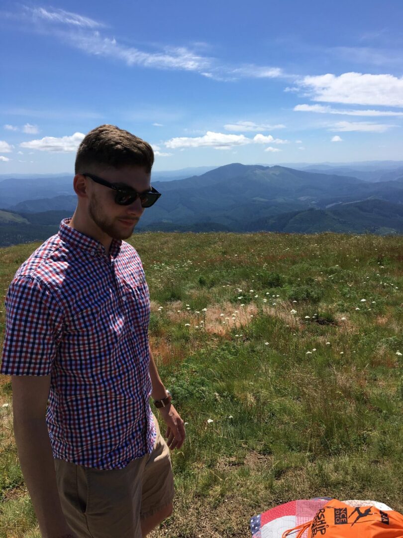 A man in sunglasses standing on a grassy hill with mountains in the background.