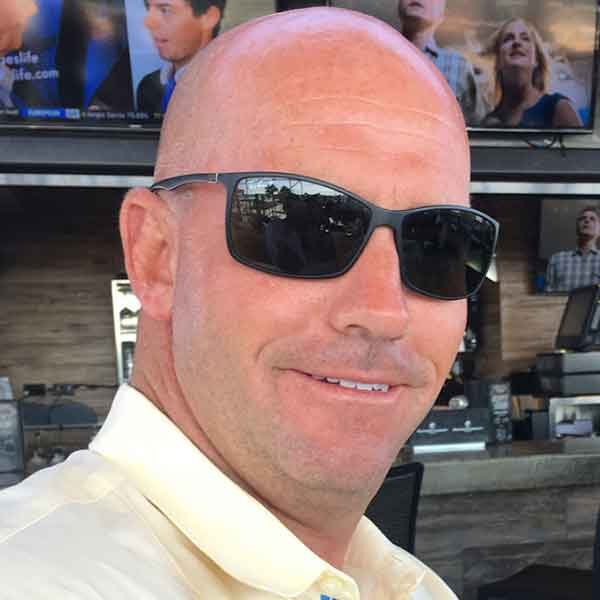 Man wearing sunglasses and a light shirt, smiling outdoors.