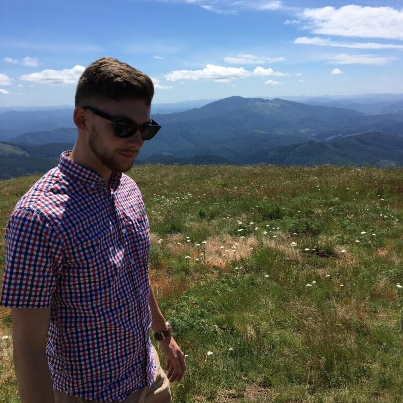 A man in sunglasses standing on a grassy hill with mountains in the background.